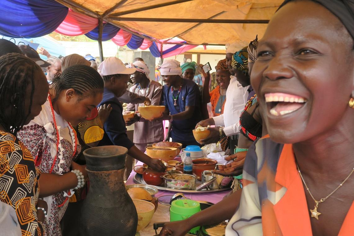 A woman is smiling as she sits in front of a table full of food. There are many other women sitting at the long table which is covered with a tent. 