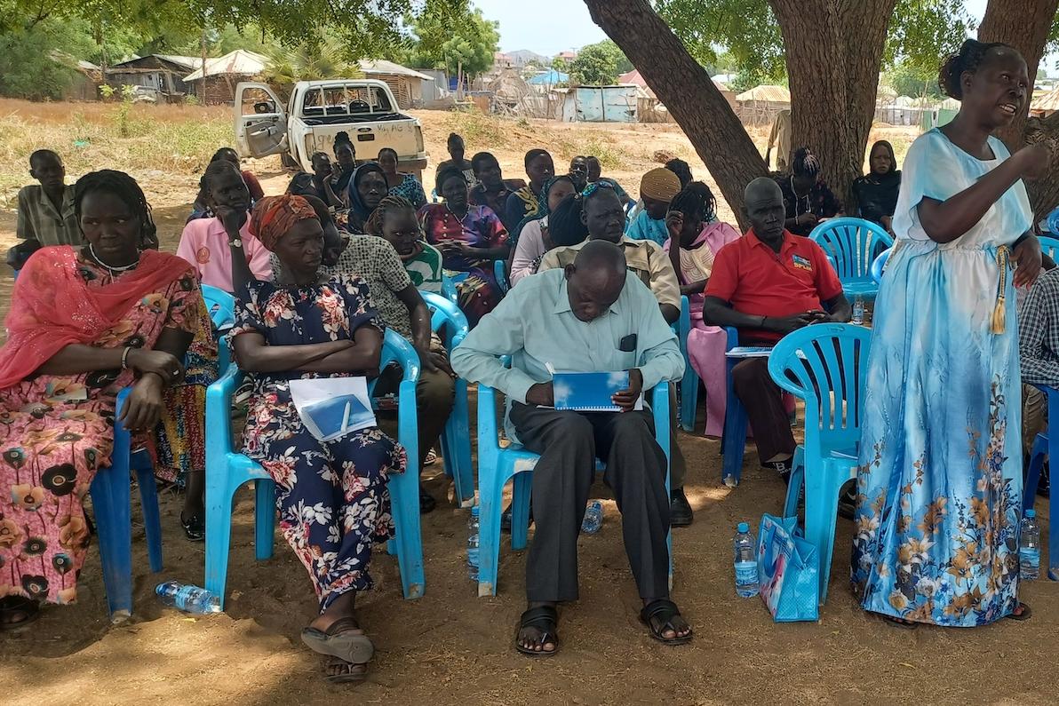 A group of people sitting in blue plastic chairs underneath a tree. In the background, there is a car with its door open.