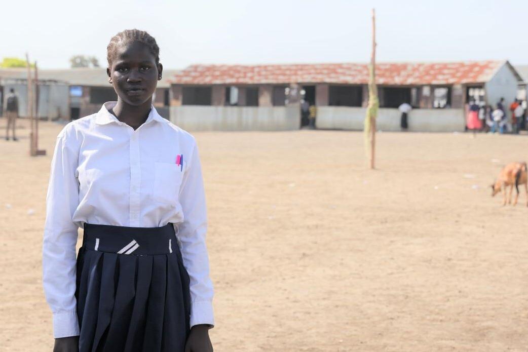 A student wearing a white shirt and dark pleated skirt stands on a dirt schoolyard with classrooms in the background and a goat grazing nearby