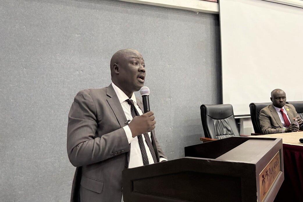 A person in a suit speaks at a podium with a microphone in a conference room, while another person sits at a table with documents and a microphone in front.