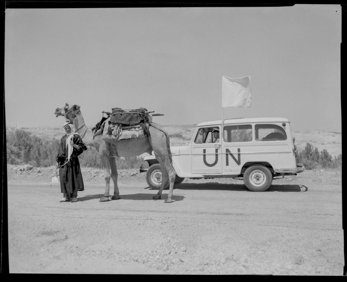 A bedoin man and his camel stand next to a UN car in the desert